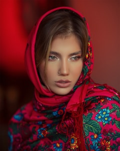 A person is wearing a vibrant, floral-patterned scarf with a red base and colorful flowers in blue, green, and yellow. The scarf is draped around the head, accentuating the person's features. The background is a warm shade of red, enhancing the vivid colors of the scarf.