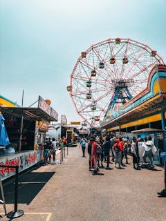 A vibrant carnival scene with a large Ferris wheel in the background. Numerous people are gathered around various food stalls and ticket booths. There is a lively atmosphere with bright colors and detailed signage on the stands.