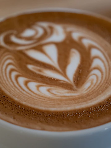 Close-up photo of a barista carefully pouring latte art with creamy milk foam in warm brown tones.