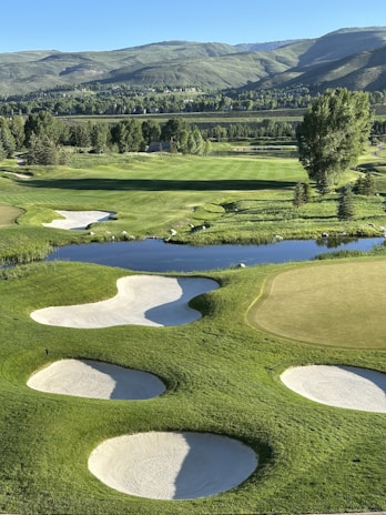 A scenic golf course with well-maintained greens and sand bunkers, surrounded by lush green trees and hills in the background. A small body of water is visible, reflecting the clear blue sky. The landscape is expansive and open, creating a serene and tranquil environment.