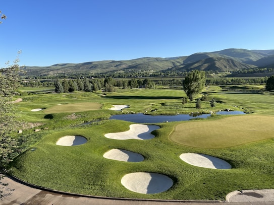 An expansive golf course set in a lush green valley with rolling hills and mountains in the background. The image captures a well-maintained green with several sand bunkers around it. There are numerous trees scattered throughout the scene, as well as small water features that add to the scenic beauty. The sky is clear and blue, indicating a bright, sunny day.