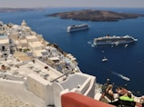 A coastal Mediterranean town features whitewashed buildings with domed roofs cascading down a hillside. Two large cruise ships are anchored in the deep blue waters, with small boats scattered around. A couple is seated on a terrace overlooking the ocean, enjoying a meal or drink under the clear sky.