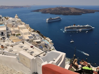 A coastal Mediterranean town features whitewashed buildings with domed roofs cascading down a hillside. Two large cruise ships are anchored in the deep blue waters, with small boats scattered around. A couple is seated on a terrace overlooking the ocean, enjoying a meal or drink under the clear sky.