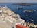 A coastal Mediterranean town features whitewashed buildings with domed roofs cascading down a hillside. Two large cruise ships are anchored in the deep blue waters, with small boats scattered around. A couple is seated on a terrace overlooking the ocean, enjoying a meal or drink under the clear sky.