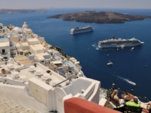 A coastal Mediterranean town features whitewashed buildings with domed roofs cascading down a hillside. Two large cruise ships are anchored in the deep blue waters, with small boats scattered around. A couple is seated on a terrace overlooking the ocean, enjoying a meal or drink under the clear sky.