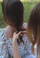 Two individuals are involved in braiding hair, with one person sitting while the other uses their hands to style the hair. A flowery pattern is visible on the seated person's clothing, and the setting appears to be outdoors with grass and greenery in the background.