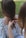 Two individuals are involved in braiding hair, with one person sitting while the other uses their hands to style the hair. A flowery pattern is visible on the seated person's clothing, and the setting appears to be outdoors with grass and greenery in the background.