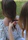 Two individuals are involved in braiding hair, with one person sitting while the other uses their hands to style the hair. A flowery pattern is visible on the seated person's clothing, and the setting appears to be outdoors with grass and greenery in the background.