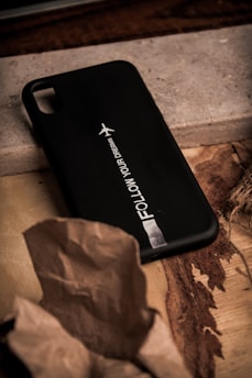 A black phone case with the text 'FOLLOW YOUR DREAMS' printed on it along with a small airplane icon. The case is placed on a textured wooden surface, partially covered by a piece of wrinkled brown paper.