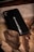 A black phone case with the text 'FOLLOW YOUR DREAMS' printed on it along with a small airplane icon. The case is placed on a textured wooden surface, partially covered by a piece of wrinkled brown paper.
