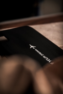 A sleek black surface with a white airplane logo and the words 'YOUR DREAMS' printed in a minimalist font. The lighting is dim, creating a subtle play of shadows on the textured background.