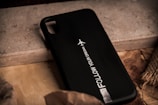 A black phone case with the text 'FOLLOW YOUR DREAMS' alongside a small graphic of an airplane. It rests on a textured surface with mixed materials like wood and concrete, surrounded by earthy tones.
