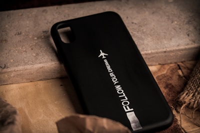 A black phone case with the text 'FOLLOW YOUR DREAMS' alongside a small graphic of an airplane. It rests on a textured surface with mixed materials like wood and concrete, surrounded by earthy tones.