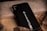 A black phone case with the text 'FOLLOW YOUR DREAMS' alongside a small graphic of an airplane. It rests on a textured surface with mixed materials like wood and concrete, surrounded by earthy tones.
