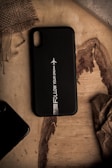 A smartphone case with the phrase 'Follow Your Dreams' and an airplane icon is placed on a wooden surface. The surrounding area has rustic elements, including burlap fabric and weathered wood. There's crumpled paper and part of another phone case visible.
