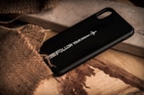 A black phone case with the text 'Follow Your Dreams' and an airplane graphic is placed on a rustic wooden surface. Part of the scene is covered with a piece of rough burlap fabric, adding texture to the composition.