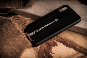 A black phone case with the text 'Follow Your Dreams' and an airplane graphic is placed on a rustic wooden surface. Part of the scene is covered with a piece of rough burlap fabric, adding texture to the composition.