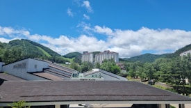 A picturesque landscape with rolling green hills under a bright blue sky with scattered white clouds. In the foreground, there are buildings with sloped roofs, and in the distance, a tall residential or hotel building is surrounded by lush greenery.