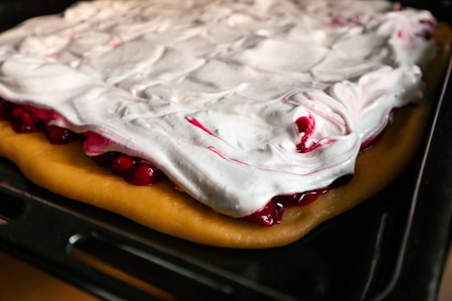 A baking tray holds a partially baked dessert featuring a thick layer of golden brown dough topped with fluffy white cream or meringue. The white cream has streaks of red filling visible underneath, adding a contrasting color and texture.