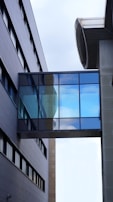 A modern architectural scene with two parallel buildings connected by a glass skybridge. The facade of the buildings features sleek, dark panels and numerous windows. The skybridge has reflective glass panels that show a glimpse of the sky above, featuring some clouds.
