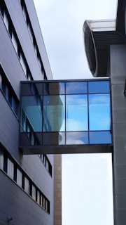 A modern architectural scene with two parallel buildings connected by a glass skybridge. The facade of the buildings features sleek, dark panels and numerous windows. The skybridge has reflective glass panels that show a glimpse of the sky above, featuring some clouds.