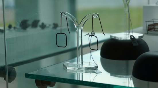 A display case containing modern, minimalist metal eyeglass frames suspended on a transparent stand. The background features a slightly blurred view, adding depth to the arrangement. The overall setting suggests a sophisticated, clean, and contemporary aesthetic.
