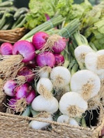 Bunches of fresh onions, both red and white, are placed in a woven basket. The onions have their long green stalks and roots still attached. The background displays more green vegetables, creating a vibrant array of produce.