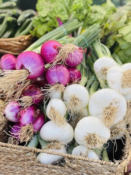 Bunches of fresh onions, both red and white, are placed in a woven basket. The onions have their long green stalks and roots still attached. The background displays more green vegetables, creating a vibrant array of produce.