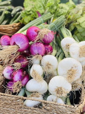 Bunches of fresh onions, both red and white, are placed in a woven basket. The onions have their long green stalks and roots still attached. The background displays more green vegetables, creating a vibrant array of produce.
