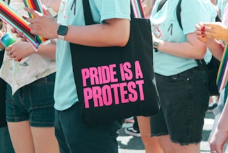 People are gathered wearing light blue shirts, some holding rainbow-colored flags or items. One person is holding a black bag with bold pink text reading 'PRIDE IS A PROTEST.'