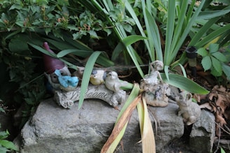 A garden setting features a collection of decorative items, including a gnome partially hidden by greenery and a selection of small bird statues in various colors perched on a stone with 'Welcome' inscribed on it. Surrounding the statues are a mix of lush green plants and a few dried brown leaves, adding contrast to the natural setting. A small stone statue resembling a rabbit is also present.