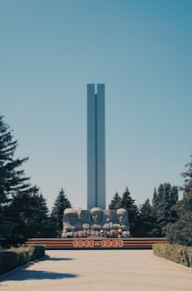 A tall, rectangular monument stands prominently against a clear blue sky, surrounded by lush evergreen trees. Below the monument, three large stone busts commemorate historical figures, accompanied by colorful floral arrangements. The years 1941-1945 are prominently displayed in bold red numerals at the base.