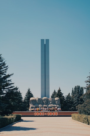 A tall, rectangular monument stands prominently against a clear blue sky, surrounded by lush evergreen trees. Below the monument, three large stone busts commemorate historical figures, accompanied by colorful floral arrangements. The years 1941-1945 are prominently displayed in bold red numerals at the base.