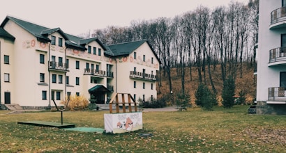 A multi-story residential building with several balconies is set against a backdrop of leafless trees, suggesting late autumn. The lawn in front of the building is scattered with fallen leaves, and there are small evergreen trees planted along the edge. A sign with cartoon graphics is positioned on the grass, adjacent to a simple children's play structure.