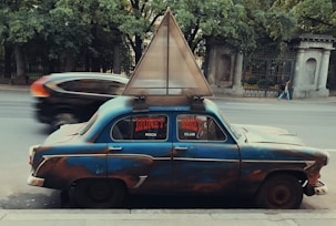 A weathered vintage car is parked on the side of a city street, featuring a unique triangular structure mounted on its roof. The windows display signage reading 'Money Rock Club' in bold red letters. A vehicle speeds by on the street, creating a blurred effect. Lush green trees and ornate stone structures form the background.