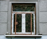 A window with an ornate iron grille featuring intricate scrollwork designs. The window is set in a textured gray wall with visible panels and a small ledge. The window frame is wooden, contrasting with the iron features.
