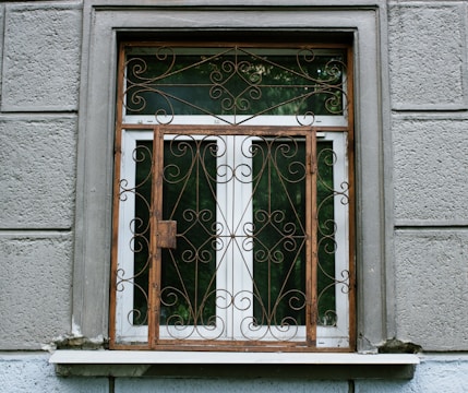 A window with an ornate iron grille featuring intricate scrollwork designs. The window is set in a textured gray wall with visible panels and a small ledge. The window frame is wooden, contrasting with the iron features.