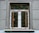 A window with an ornate iron grille featuring intricate scrollwork designs. The window is set in a textured gray wall with visible panels and a small ledge. The window frame is wooden, contrasting with the iron features.