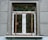 A window with an ornate iron grille featuring intricate scrollwork designs. The window is set in a textured gray wall with visible panels and a small ledge. The window frame is wooden, contrasting with the iron features.