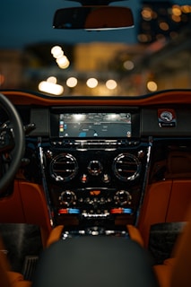The interior of a luxury car is shown, featuring a high-tech dashboard with a central screen displaying navigation instructions. The dashboard has shiny chrome accents and controls for air conditioning. The image is shot at night, with blurred city lights visible through the windshield.