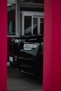 A luxurious black car is partially visible, framed by red pillars, with large glass windows and a modern building facade in the background.