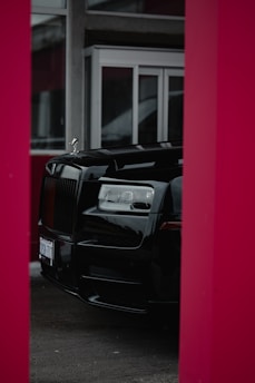 A luxurious black car is partially visible, framed by red pillars, with large glass windows and a modern building facade in the background.
