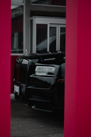 A luxurious black car is partially visible, framed by red pillars, with large glass windows and a modern building facade in the background.