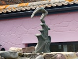 A sculpture of an abstract form is positioned in front of a pink, textured wall. The sculpture resembles a hand emerging from three curling waves, crafted from a dark, speckled material. Surrounding the base are several stones and rocks, adding to the overall earthy composition.