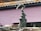 A sculpture of an abstract form is positioned in front of a pink, textured wall. The sculpture resembles a hand emerging from three curling waves, crafted from a dark, speckled material. Surrounding the base are several stones and rocks, adding to the overall earthy composition.