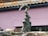 A sculpture of an abstract form is positioned in front of a pink, textured wall. The sculpture resembles a hand emerging from three curling waves, crafted from a dark, speckled material. Surrounding the base are several stones and rocks, adding to the overall earthy composition.
