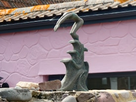 A sculpture of an abstract form is positioned in front of a pink, textured wall. The sculpture resembles a hand emerging from three curling waves, crafted from a dark, speckled material. Surrounding the base are several stones and rocks, adding to the overall earthy composition.