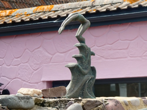 A sculpture of an abstract form is positioned in front of a pink, textured wall. The sculpture resembles a hand emerging from three curling waves, crafted from a dark, speckled material. Surrounding the base are several stones and rocks, adding to the overall earthy composition.