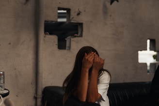 A person with long hair covers their face with their hands while sitting on a leather sofa. The background shows an unfinished and slightly damaged wall with holes revealing some structural elements. A can of beverage is placed on a surface to the left.