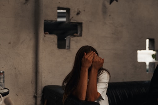 A person with long hair covers their face with their hands while sitting on a leather sofa. The background shows an unfinished and slightly damaged wall with holes revealing some structural elements. A can of beverage is placed on a surface to the left.
